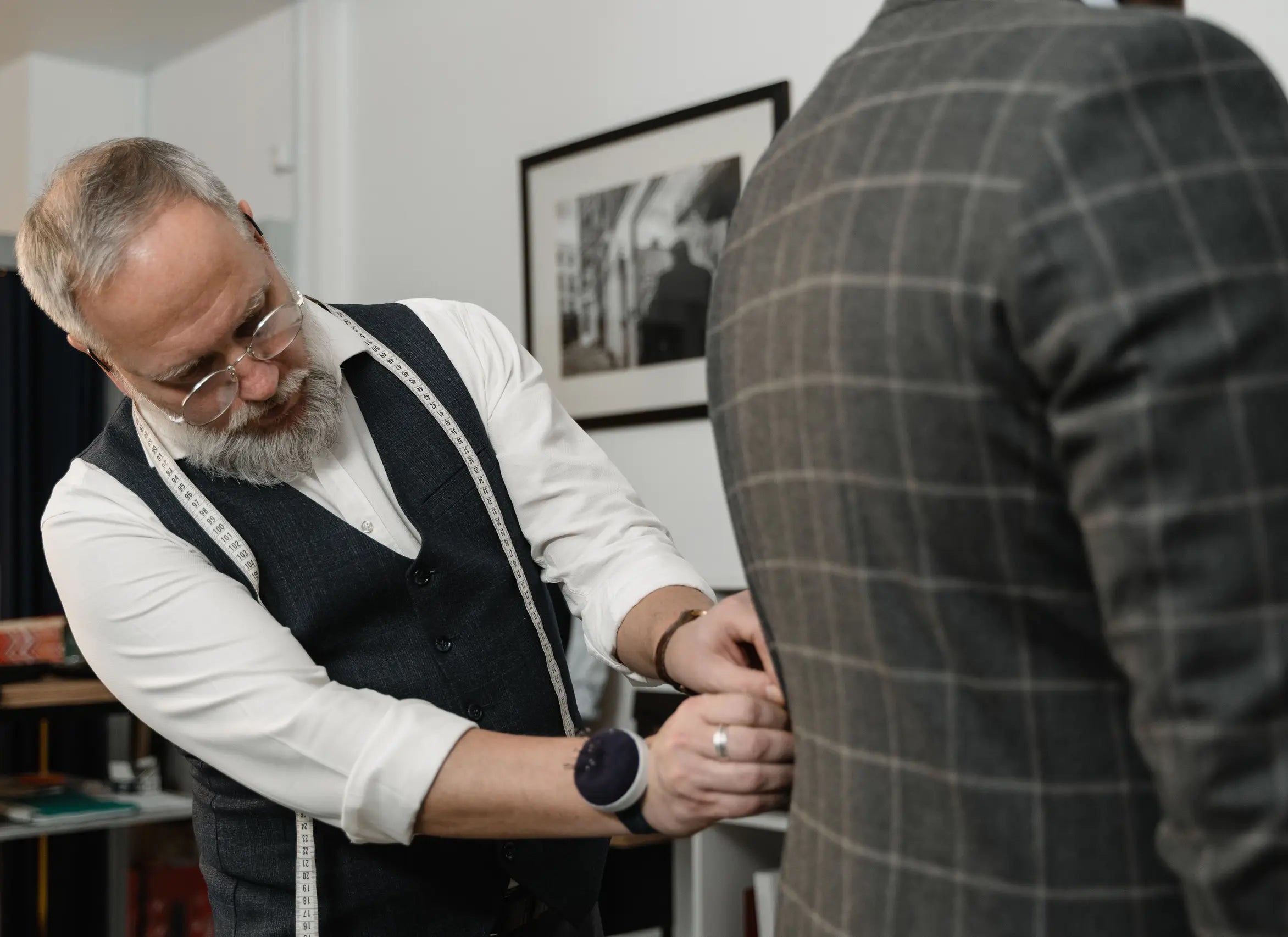 Tailor with glasses and vest adjusting a client's grey checkered suit jacket indoors