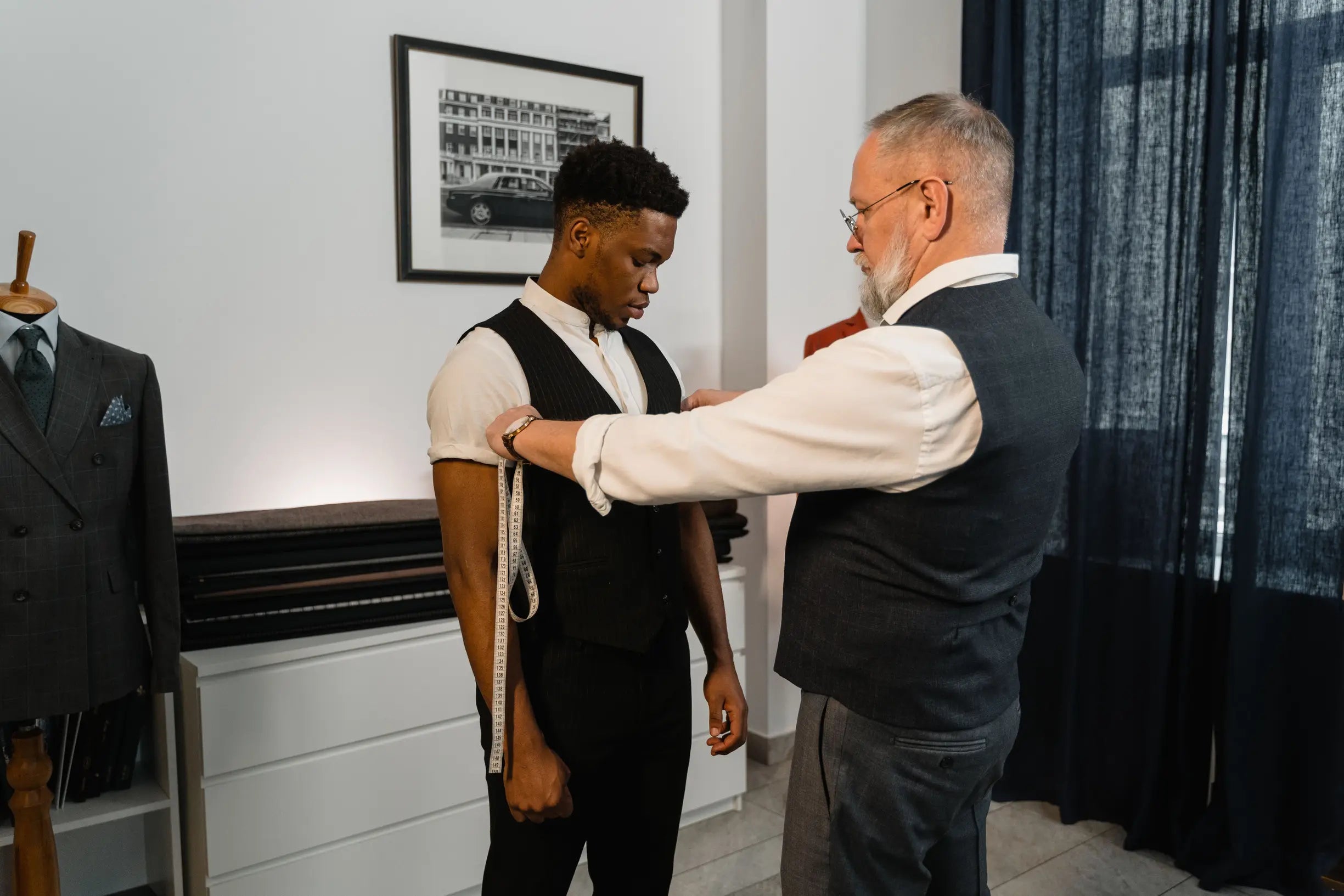 Older man measuring a younger man’s arm length wearing a dark tailored vest and white shirt indoors