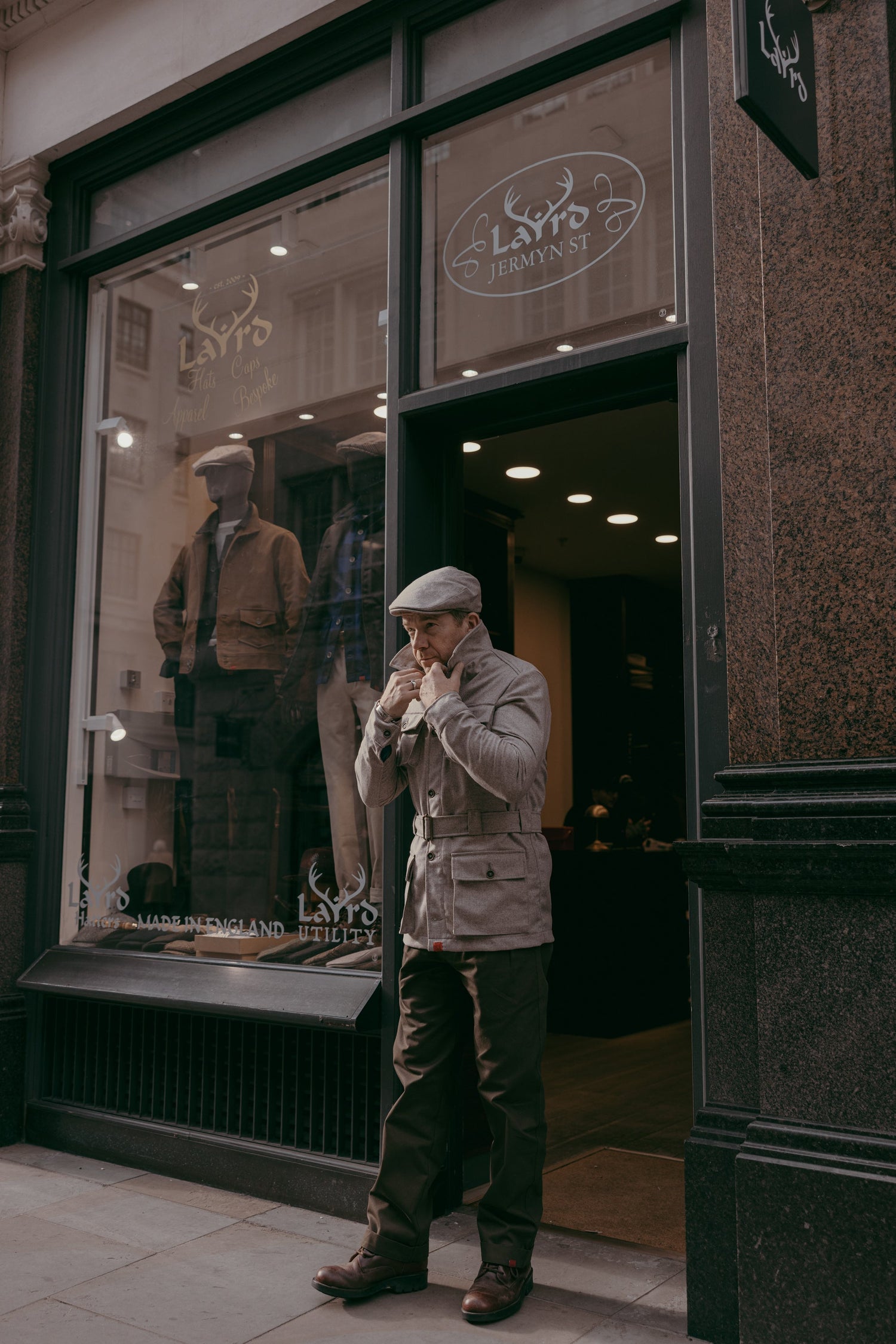 Man adjusting collar of beige belted jacket with flat cap outside Laird Utility store on Jermyn Street