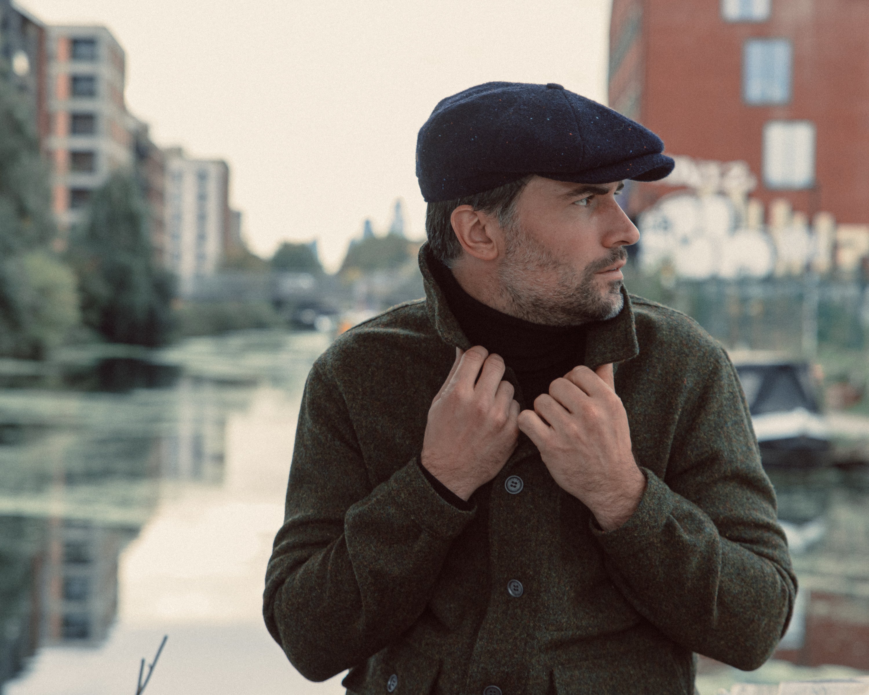 Man outdoors by water wearing dark green buttoned coat and dark flat cap, holding up coat collar, looking right