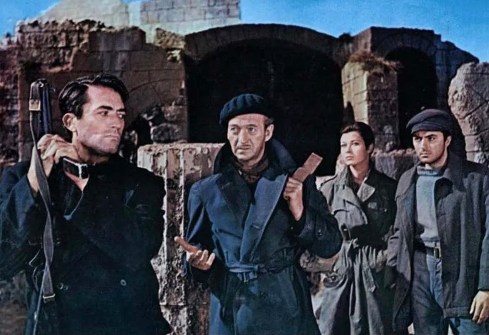 Group of four in dark coats and berets standing before ancient stone ruins under blue sky