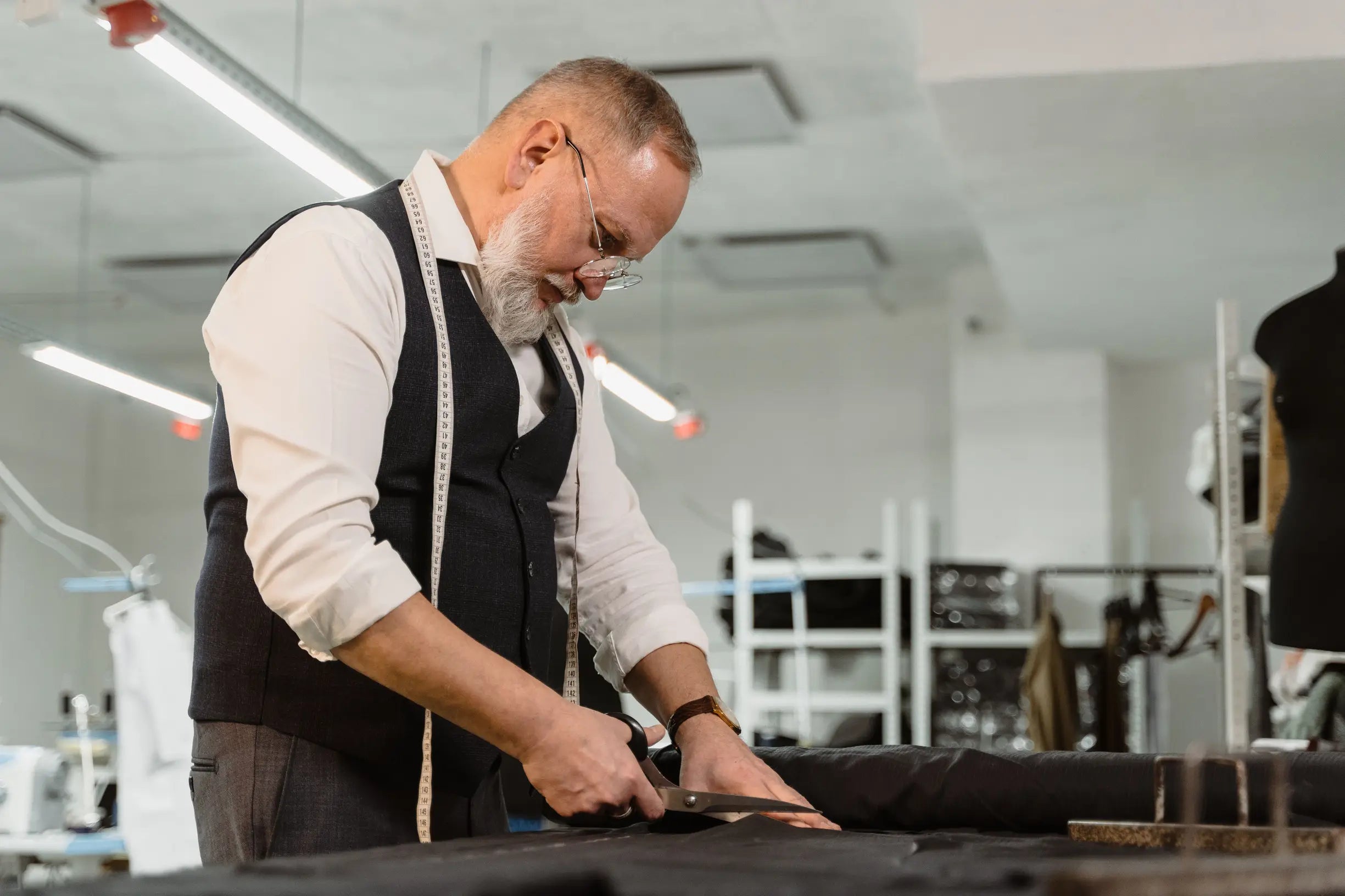 Older man with gray beard cutting black fabric wearing white shirt and dark vest in tailoring workshop