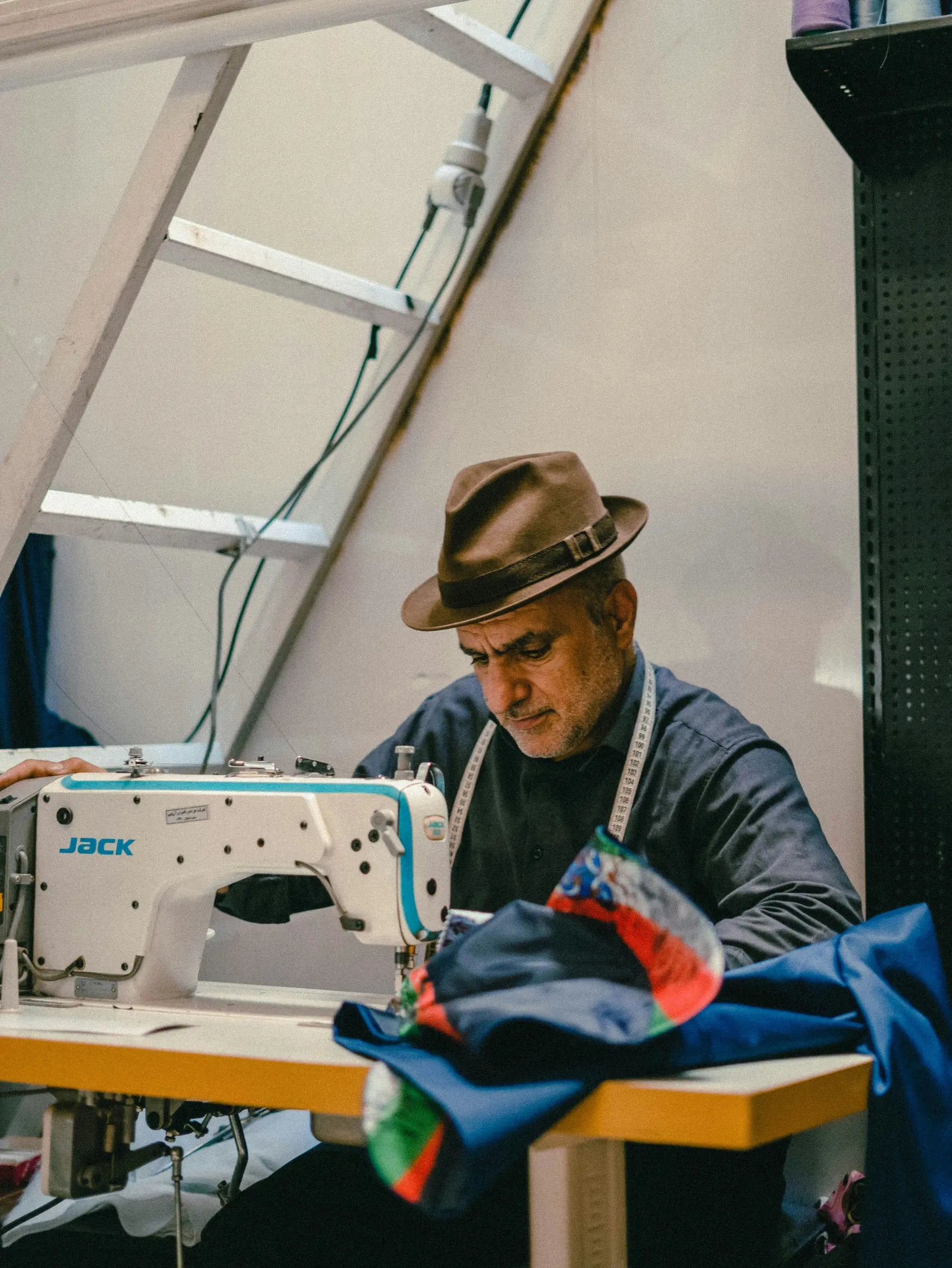 Older man wearing a brown hat and dark shirt sewing colourful fabric on an industrial machine at eye level