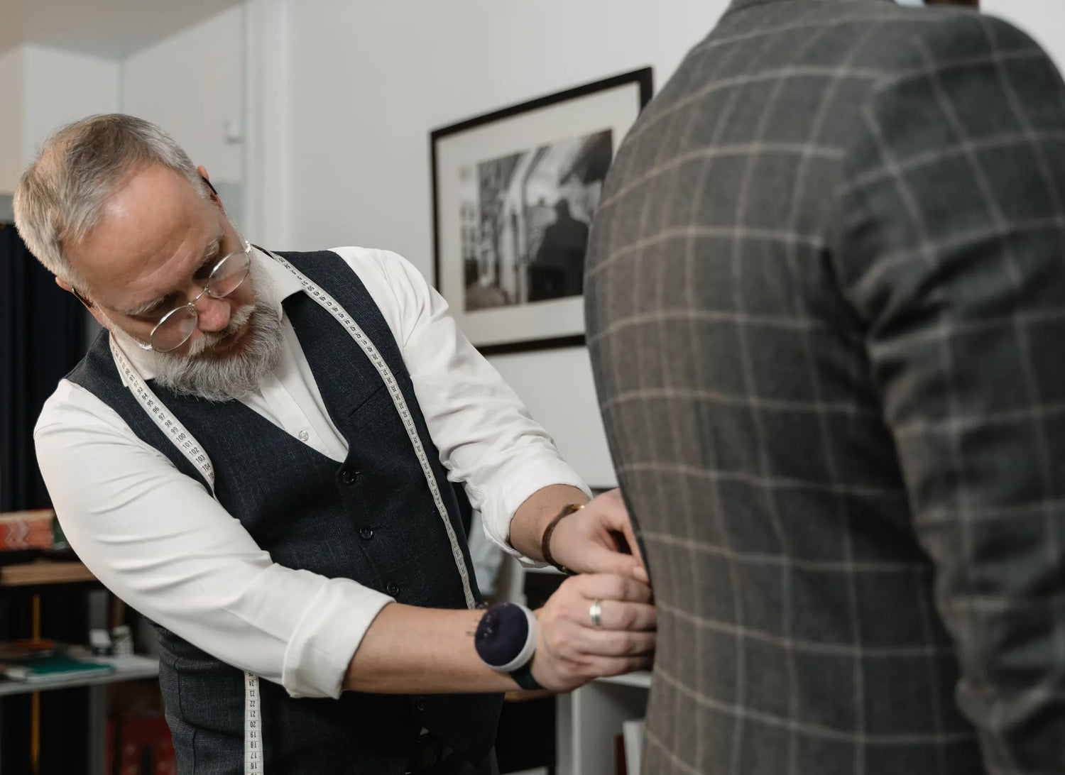Tailor with glasses and vest adjusting a client's grey checkered suit jacket indoors
