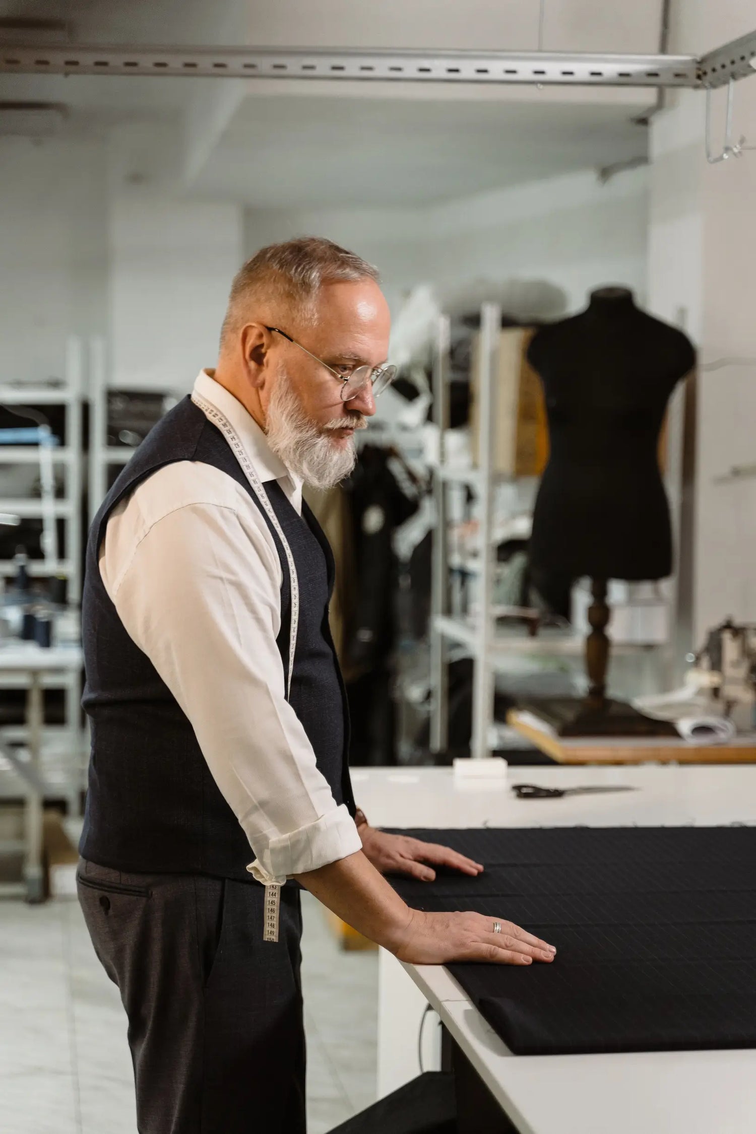 Older man inspects black fabric on table in tailoring studio with measuring tape around neck