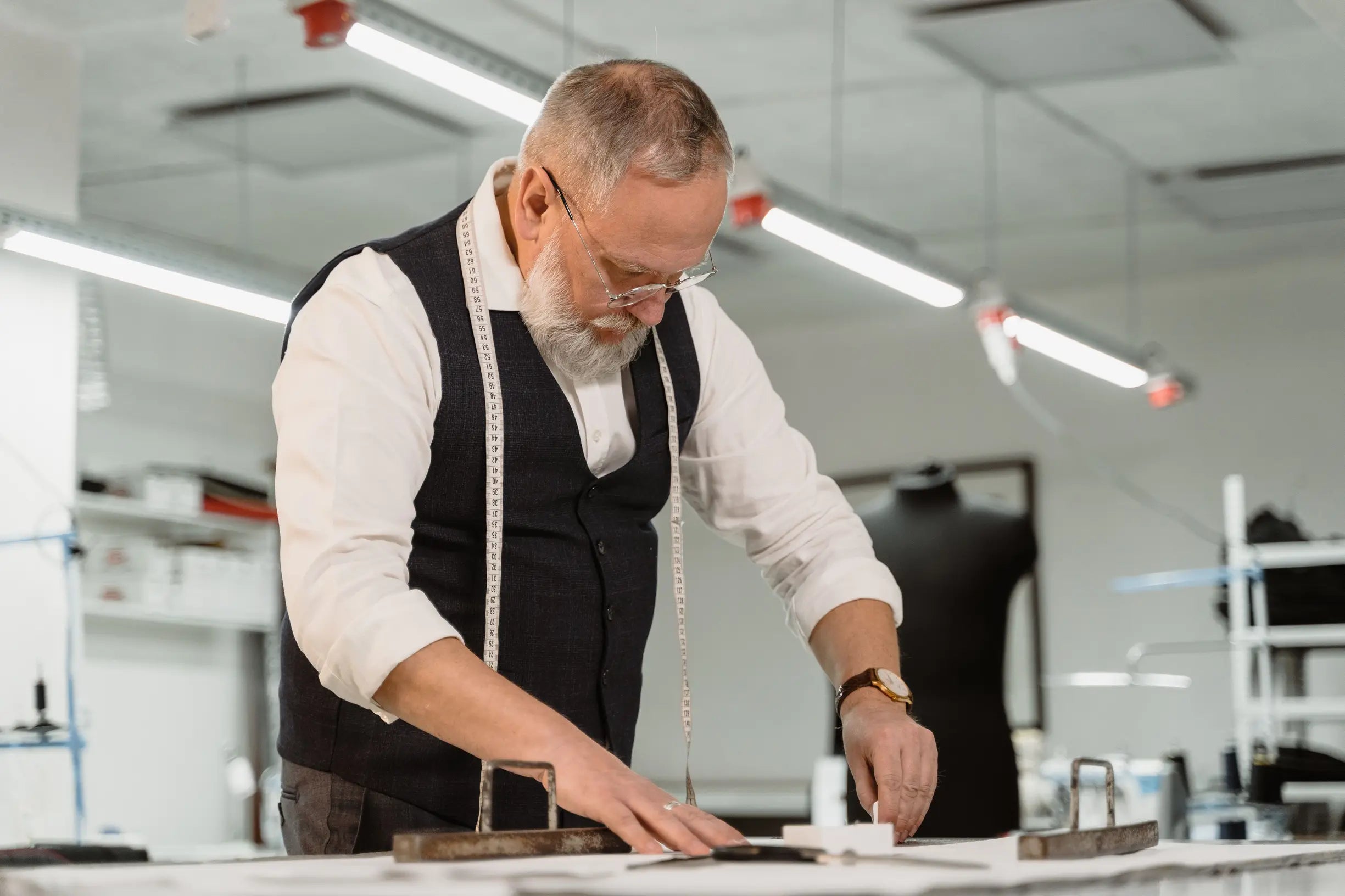 Older man in white shirt and dark waistcoat working with patterns on a table in a bright tailoring workshop