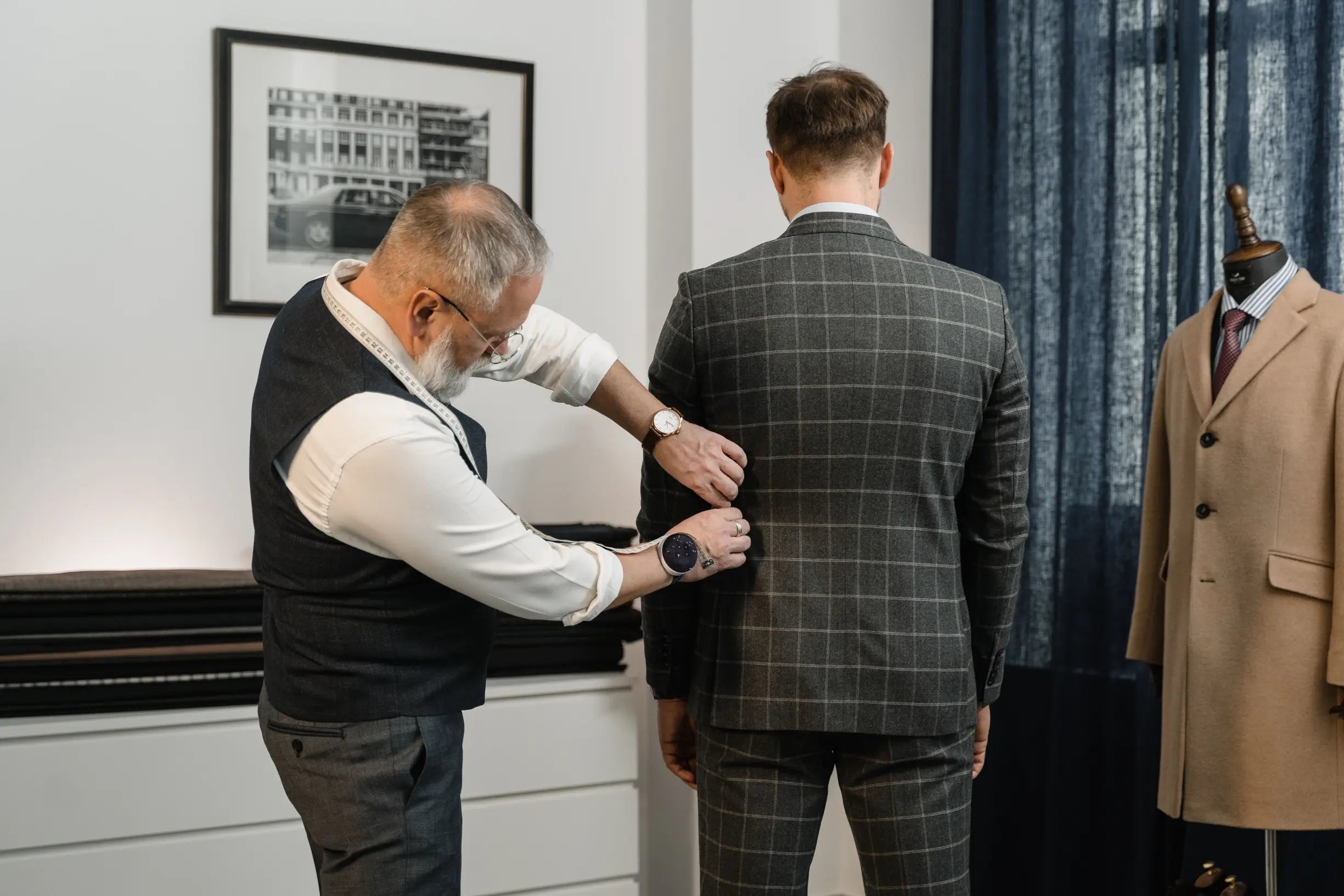 Tailor adjusting the sleeve of a gray checkered suit on a customer from behind, with a beige overcoat on a mannequin nearby
